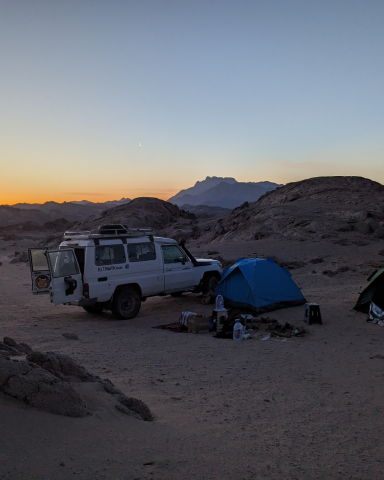 Ein Jeep steht neben einem blauen Zelt in der Wüste bei Sonnenuntergang.