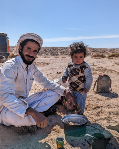 Ein Mann und ein Kind sitzen im Sand, neben einem Eimer, mit Wüstenlandschaft im Hintergrund.